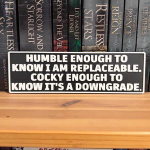 May include: A rectangular black sign with a white border and white embossed text that reads: HUMBLE ENOUGH TO KNOW I AM REPLACEABLE. COCKY ENOUGH TO KNOW IT'S A DOWNGRADE. The sign features a clean, bold font and a high-contrast design.