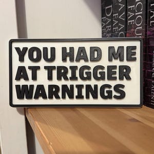 Könnte beinhalten: Ein rechteckiges Schild mit dem Text "YOU HAD ME AT TRIGGER WARNINGS" in schwarzen, erhabenen Buchstaben. Das Schild hat einen weißen Hintergrund und einen schwarzen Rand. Es wird auf einem Holzregal mit Büchern im Hintergrund angezeigt.