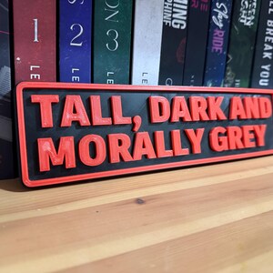 May include: A rectangular sign with the text "TALL, DARK AND MORALLY GREY" in red, raised lettering. The sign has a black background and a red border. The sign is placed in front of a row of books.