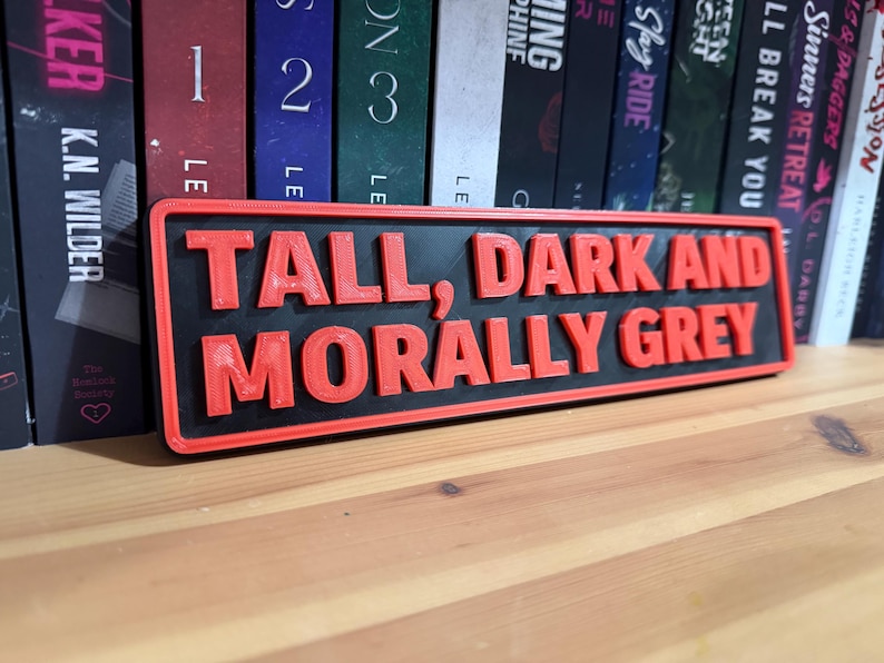 May include: A rectangular sign with the text "TALL, DARK AND MORALLY GREY" in red, raised lettering. The sign has a black background and a red border. The sign is placed in front of a row of books.