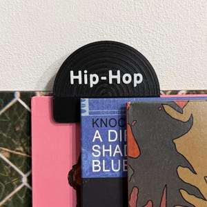 May include: A black vinyl record-shaped bookmark with the white text "Hip-Hop" on top of a stack of books. The books have pink, blue, and brown covers. The bookmark is a unique accessory for music lovers and book enthusiasts.