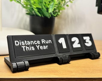 May include: Black 3D-printed distance tracker with white text reading "Distance Run This Year" and numbered tiles 1, 2, and 3. The tracker is on a wooden surface with a potted plant in the background.