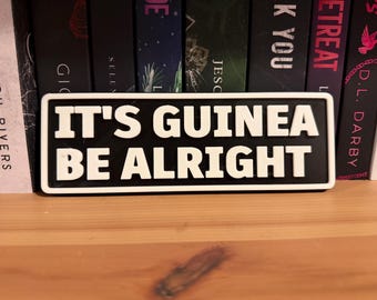 May include: A black and white rectangular sign with the text "IT'S GUINEA BE ALRIGHT" in bold white letters. The sign is placed in front of a row of books on a wooden shelf.