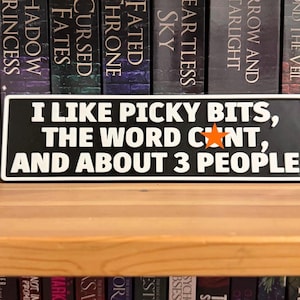 May include: A black and white rectangular sign with the text "I LIKE PICKY BITS, THE WORD CUNT, AND ABOUT 3 PEOPLE" in white lettering. An orange star is between "WORD" and "CUNT". The sign is on a wooden shelf in front of a row of books.