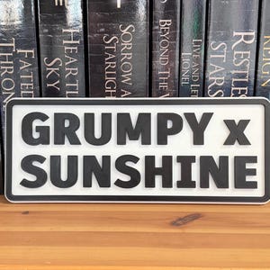 May include: A rectangular sign with the words "GRUMPY X SUNSHINE" in bold, black letters. The sign has a white background and a black border. The sign is displayed on a wooden surface in front of a row of books.