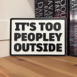 Könnte beinhalten: Rechteckiges Schild mit dem Text „IT'S TOO PEOPLEY OUTSIDE“ in schwarzen, erhabenen Buchstaben auf weißem Grund. Das Schild hat einen schwarzen Rahmen und steht auf einer Holzoberfläche, mit Büchern im Hintergrund.