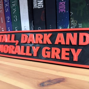 May include: A rectangular sign with the text "TALL, DARK AND MORALLY GREY" in red, raised lettering. The sign has a black background and a red border, placed on a wooden surface.