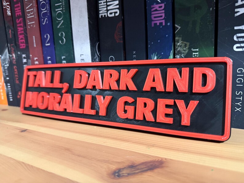 May include: A rectangular sign with the text "TALL, DARK AND MORALLY GREY" in red, raised lettering. The sign has a black background and a red border, placed on a wooden surface.
