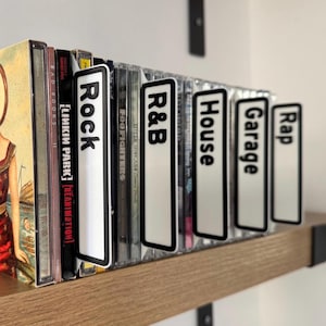 May include: A collection of CDs on a wooden shelf, with genre dividers labeled "Rock", "R&B", "House", "Garage", and "Rap". The dividers are white with black borders and text. The shelf is mounted on a white wall.