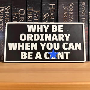 May include: A rectangular black sign with white lettering that reads "WHY BE ORDINARY WHEN YOU CAN BE A C*NT" with a blue star replacing the "O". The sign has a white border and is displayed in front of a bookshelf.