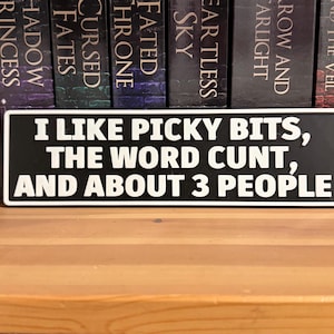 Könnte beinhalten: Ein rechteckiges Schild in Schwarz und Weiß mit dem Text "I LIKE PICKY BITS, THE WORD CUNT, AND ABOUT 3 PEOPLE". Das Schild befindet sich vor einem Bücherregal.