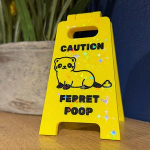 May include: A bright yellow caution sign with a cartoon ferret illustration and the words "CAUTION" and "FERRET POOP". The sign has a handle at the top and is decorated with holographic starbursts.