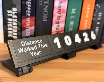May include: A black 3D-printed counter displaying "Distance Walked This Year" with the number "10426" in white. The counter is on a wooden surface, with a row of books in the background.