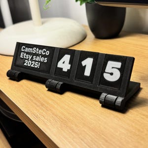May include: A black Etsy sales counter with white numbers. The counter displays "415" and the text "CamSteCo Etsy sales 2025!" on a wooden surface. The counter has a flip design.
