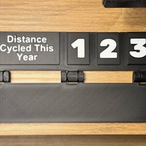 May include: A black 3D-printed cycling distance tracker with the text "Distance Cycled This Year" and the numbers "123" in white. The tracker is mounted on a wooden surface and has a hinged design.