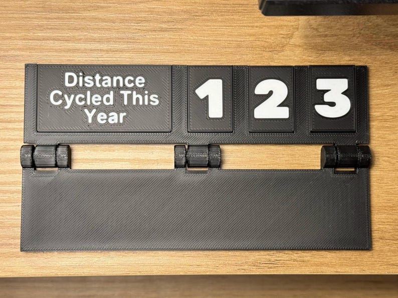 May include: A black 3D-printed cycling distance tracker with the text "Distance Cycled This Year" and the numbers "123" in white. The tracker is mounted on a wooden surface and has a hinged design.