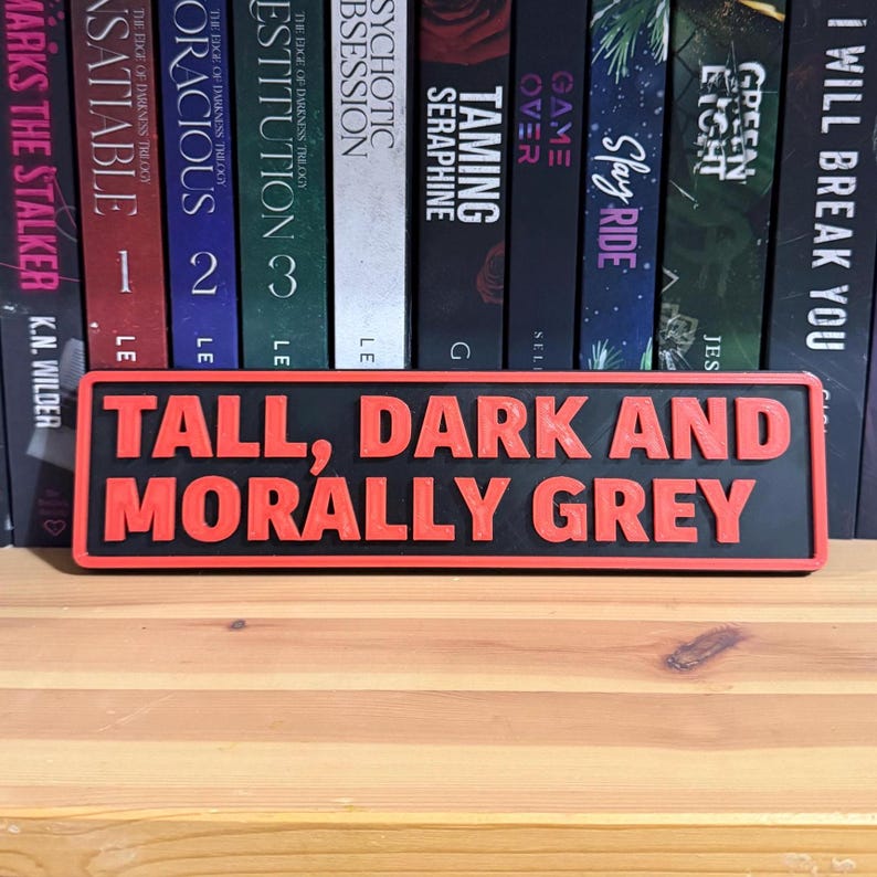 May include: A black rectangular sign with the words "TALL, DARK AND MORALLY GREY" in red, raised lettering. The sign has a red border and is displayed in front of a bookshelf.