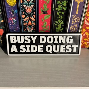 May include: A black and white sign with the text "BUSY DOING A SIDE QUEST". The sign is rectangular and has a white border. The sign is in front of a collection of colourful bookmarks.