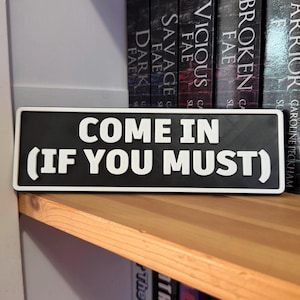 Könnte beinhalten: Ein rechteckiges schwarz-weißes Schild mit dem Text "COME IN (IF YOU MUST)". Das Schild befindet sich auf einem Holzregal, im Hintergrund sind Buchrücken zu sehen.