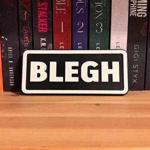 May include: A rectangular black and white sign with the word "BLEGH" in bold, white letters. The sign is set against a backdrop of colorful book spines on a wooden shelf.