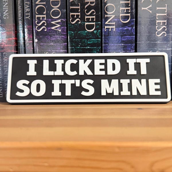 May include: A black and white rectangular sign with the text "I LICKED IT SO IT'S MINE" in bold, white letters. The sign is placed in front of a row of books on a wooden shelf.