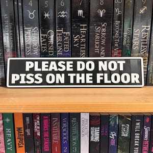 Puede incluir: Un letrero en blanco y negro con el texto "PLEASE DO NOT PISS ON THE FLOOR". El letrero rectangular está colocado en una estantería de madera frente a una colección de libros.