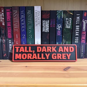 May include: A black and red rectangular sign with the text "TALL, DARK AND MORALLY GREY" in red. The sign is in front of a bookshelf filled with books.