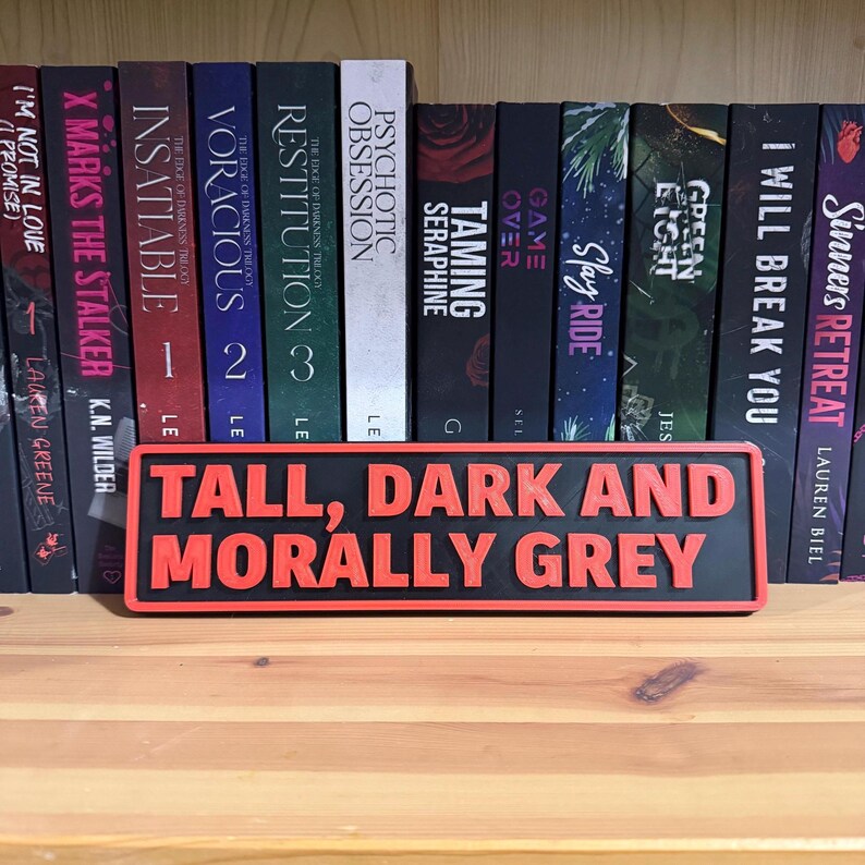May include: A black and red rectangular sign with the text "TALL, DARK AND MORALLY GREY" in red. The sign is in front of a bookshelf filled with books.