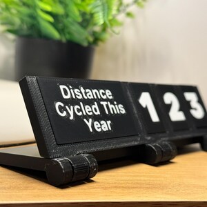 May include: A black 3D-printed desk calendar displaying "Distance Cycled This Year" with the numbers "123". The calendar is on a wooden surface, with a green plant in a black pot in the background.
