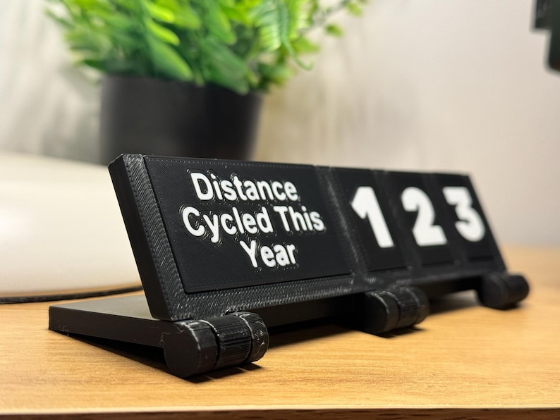 May include: A black 3D-printed desk calendar displaying "Distance Cycled This Year" with the numbers "123". The calendar is on a wooden surface, with a green plant in a black pot in the background.