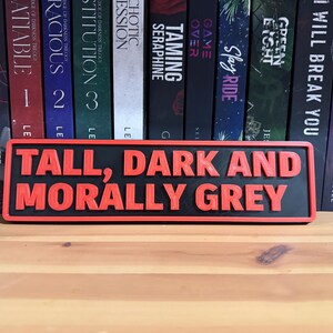 May include: A black and red rectangular sign with the words "TALL, DARK AND MORALLY GREY" in raised, red letters. The sign is displayed in front of a shelf of books.
