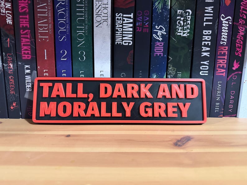 May include: A black and red rectangular sign with the words "TALL, DARK AND MORALLY GREY" in raised, red letters. The sign is displayed in front of a shelf of books.