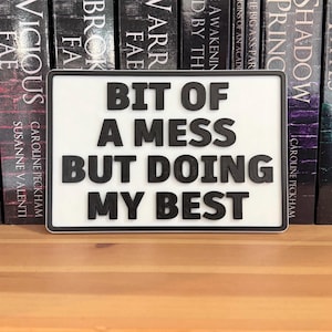 May include: A white rectangular sign with black text saying "BIT OF A MESS BUT DOING MY BEST." The sign has a black border and is placed in front of a row of books.