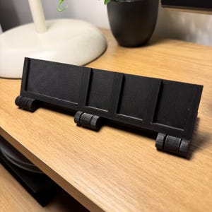 May include: A black, rectangular desk organizer with four sections and two sets of cylindrical feet. The organizer is made of a matte black material and sits on a light brown wooden surface. The design is simple and modern.