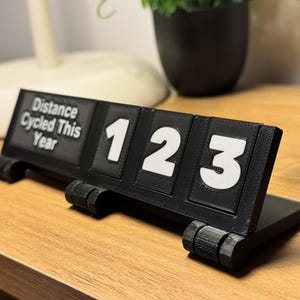May include: A black 3D-printed cycling distance counter with white numbers 123. The text "Distance Cycled This Year" is printed on the left side. The counter is on a wooden surface.