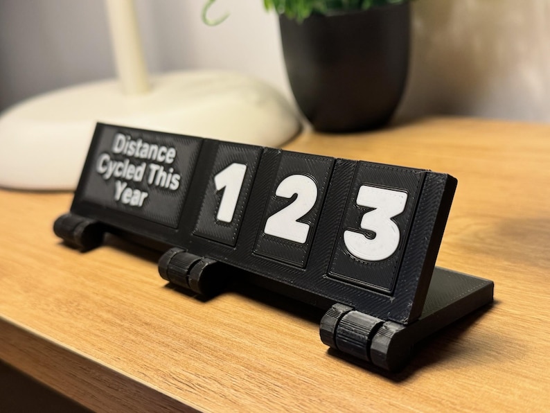 May include: A black 3D-printed cycling distance counter with white numbers 123. The text "Distance Cycled This Year" is printed on the left side. The counter is on a wooden surface.