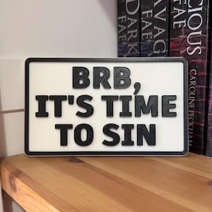 May include: A rectangular sign with a white background and a black border. The sign features the phrase "BRB, IT'S TIME TO SIN" in bold, black letters. The sign is displayed on a wooden shelf, with books in the background.
