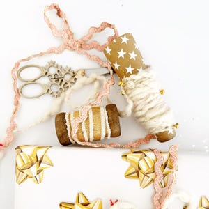 May include: A close-up of white gift wrap with gold star-shaped bows. There is a spool of gold and white ribbon, a spool of pink ribbon, and a spool of white yarn with gold beads.