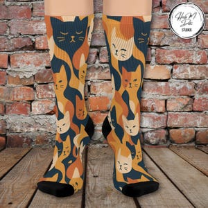 May include: A pair of knee-high socks featuring a repeating pattern of stylized cat faces in shades of orange, tan, and navy blue. The socks have black heels and toes. The background is a brick wall and wooden floor.