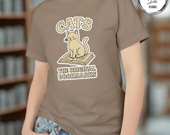 May include: A tan t-shirt featuring a cream-colored graphic of a cat sitting on a book. The text above the cat reads "CATS", and below it, "THE ORIGINAL BOOKMARKS". The shirt is a neutral color and has short sleeves.