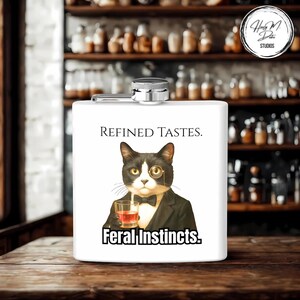 May include: White stainless steel flask with a hinged cap. The front features a tuxedo cat illustration wearing a monocle and holding a drink, with the text "REFINED TASTES." above and "Feral Instincts." below.