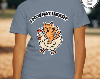 May include: A light blue t-shirt featuring a cartoon illustration of an orange tabby cat riding a white chicken. The text "I DO WHAT I WANT" is arched above the cat and chicken. The shirt is made of a soft material.