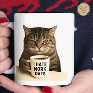 Puede incluir: Taza de cerámica blanca con un gato atigrado gruñón sosteniendo una taza de café. La taza tiene el texto "I HATE WORK DAYS" impreso. El gato tiene ojos verdes y una expresión de disgusto. La taza es sostenida por una persona con esmalte de uñas rojo.