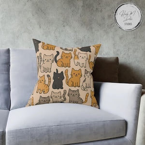 Könnte beinhalten: Quadratisches Zierkissen mit einem sich wiederholenden Muster von Cartoon-Katzen in verschiedenen Farben, darunter Orange, Grau und Schwarz, auf beigem Hintergrund. Das Kissen liegt auf einem weißen Sofa. Das Kissen hat den Text "Handy M Dake Studios" in der oberen rechten Ecke.