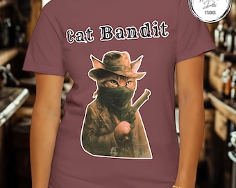 May include: A maroon t-shirt with a graphic of a cat bandit wearing a hat and a mask, holding a gun. The text "Cat Bandit" is above the graphic in a stylized font. The shirt has short sleeves.