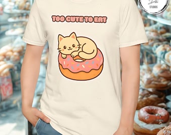 May include: Cream-colored t-shirt featuring a cartoon cat lying on a pink donut with sprinkles. The text "TOO CUTE TO EAT" is above the cat and donut graphic. The shirt is a casual style.
