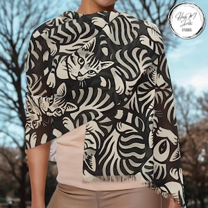 May include: A black and white patterned scarf featuring a cat design. The scarf is draped over the shoulders and has a fringe along the bottom edge. The scarf is made of a lightweight material.