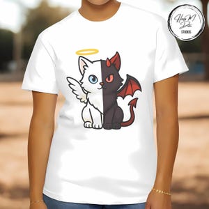 May include: White t-shirt featuring a split design of a cat, half angel and half devil. The angel side is white with a halo, wings, and a blue eye. The devil side is black with horns, a red wing, and a red eye. The shirt has a round neck.