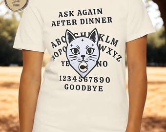 May include: Cream-colored t-shirt featuring a stylized cat design with a fish in its mouth, surrounded by letters, numbers, and the phrases "ASK AGAIN AFTER DINNER," "YE," "NO," and "GOODBYE."