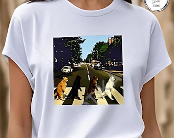 May include: White t-shirt featuring a colorful graphic of cats crossing a street, a parody of the iconic Abbey Road album cover. The image includes a road, trees, and a blue sky. The shirt has short sleeves.